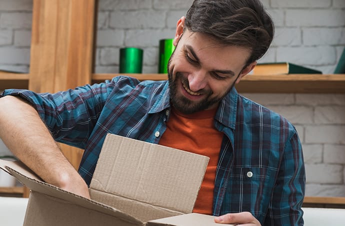 Lachende man opent een kartonnen doos en kijkt nieuwsgierig naar de inhoud, zittend in een woonkamer met boekenplanken op de achtergrond.